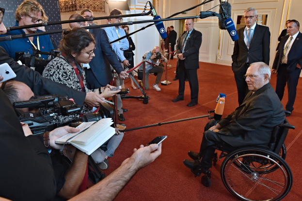 Ein Mann im Rollstuhl umgeben von Reportern mit Kameras, Handys und Mikrofonen sowie einem Banner und Scheinwerfern im Hintergrund.