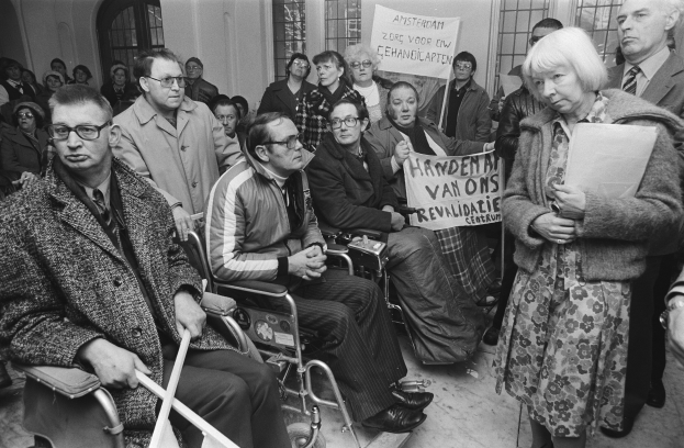 Eine Gruppe von Menschen in Rollstühlen und vor einer Menge stehend, einige halten Schilder, mit Fenstern und einer Wand im Hintergrund, in Schwarz-Weiß.