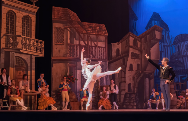 Zwei Tänzer performen auf der Bühne der Royal Opera House in London, während das Publikum sitzt und steht, mit einer projizierten Stadtkulisse im Hintergrund.