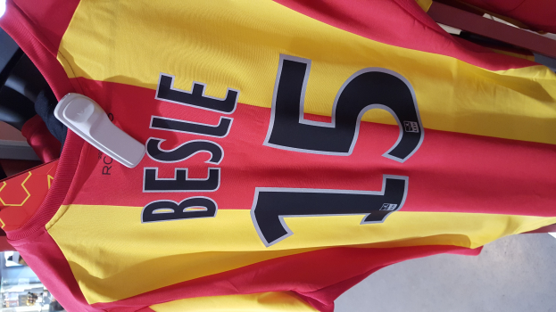 Rotes und gelbes Fußballtrikot mit Text und Nummern, das an einer Stange in einem Geschäft hängt, mit anderen Gegenständen und dem Boden im Hintergrund.