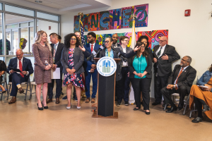 Gruppe von Menschen vor einem Rednerpult mit Mikrofon und einer Tafel mit der Aufschrift "Sancho Stadtverordnetenvorsteher tritt dem Stadtverordnetenkollegium bei", mit sitzenden und stehenden Zuh├Ârern, Flaggen, Feuerl├Âscher und Deckenleuchten im Hintergrund.