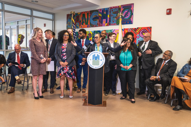 Gruppe von Menschen vor einem Rednerpult mit Mikrofon und einer Tafel mit der Aufschrift "Sancho Stadtverordnetenvorsteher tritt dem Stadtverordnetenkollegium bei", mit sitzenden und stehenden Zuh├Ârern, Flaggen, Feuerl├Âscher und Deckenleuchten im Hintergrund.