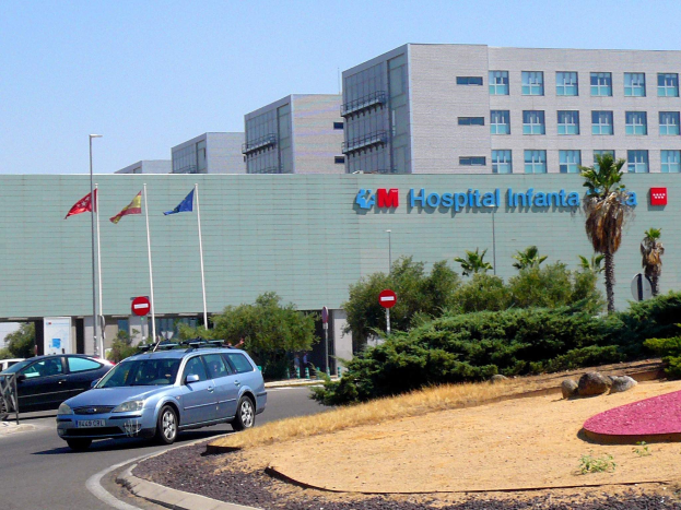 Krankenhaus mit geparkten Autos, umgeben von städtischen Elementen einschließlich Gebäuden, Straßenmöbeln, Fahrzeugen, Grünflächen und einem klaren Himmel.