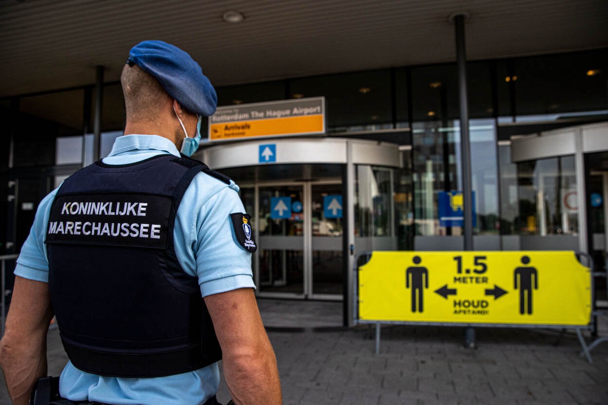 Ein Polizist in blauer Uniform und Maske steht vor einem Glasgebäude, mit gelben und anderen Informationsschildern im Hintergrund.