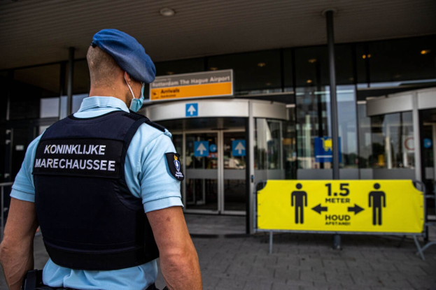 Ein Polizist in blauer Uniform und Maske steht vor einem Glasgebäude, mit gelben und anderen Informationsschildern im Hintergrund.