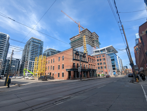 Eine belebte Straße in Toronto mit Fahrzeugen, Füpßern, Oberleitungen, Schildern und Gebäuden, mit einer Baustelle mit einem Kran im bewölktem Himmel.
