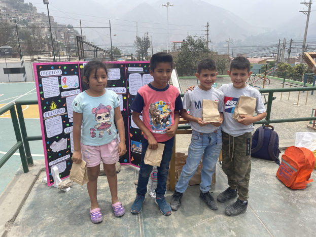 Kinder auf einem Basketballfeld mit Papierbeuteln in den Händen, mit Beuteln und Gegenständen rechts und einer Tafel mit Text links, vor dem Hintergrund von Zäunen, Pfosten, Straßenlaternen, Bäumen, Gebäuden, Hügeln und Himmel.