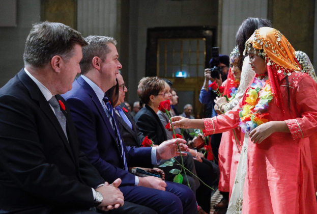 Eine Gruppe von Menschen, die in einem Raum zusammen sitzen, mit einer Frau in der Mitte, die einen Kranz trägt und Blumen hält, flankiert von einem Mann in Anzug und Krawatte rechts von ihr und einer Person mit einer Kamera links von ihr, vor einer Wand mit einer Tür.