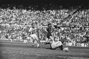 Schwarzes Foto von Fussballspielern im Einsatz auf einem Feld mit Zuschauern im Hintergrund und einer Bundesliga 1978-79 Ajax 1-1 Hannover 96-Banner.