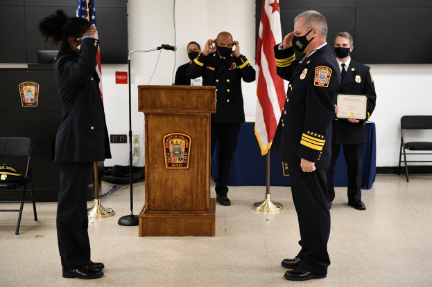 Gruppe uniformierter Personen bei einer Vereidigung eines neuen Feuerwehrchefs, der ein Zertifikat hält, mit Flaggen, Podium und maskierten Teilnehmern im Hintergrund.
