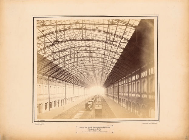 Ein altes Foto eines Bahnhofs mit einem Zug auf den Schienen, umgeben von Gebäuden mit Fenstern und einem Dach, das von Metallstreben gestützt wird, mit Text am unteren Rand des Bildes.