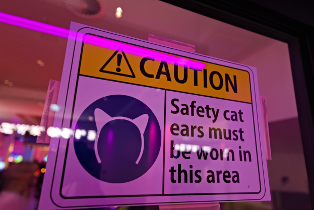 Hellgelbes Warnschild in einem Ladfenster mit fetter schwarzer Schrift, die "Vorsicht: Sicherheits-Katzenohren müssen in diesem Bereich getragen werden" lautet.