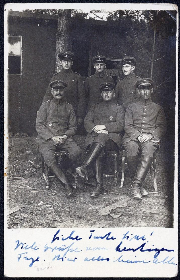 Schwarzes und weißes Foto einer Gruppe von Männern in deutschen Militäruniformen aus dem Ersten Weltkrieg, die vor einem Haus mit Bäumen im Hintergrund auf Stühlen sitzen.
