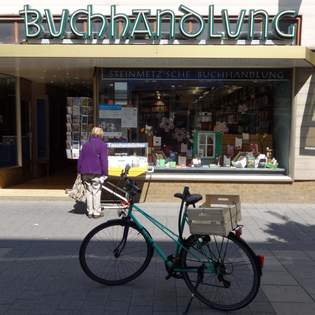 Eine Frau steht neben einem Fahrrad, das vor einem Laden geparkt ist, mit einem Fahrradrad in Vordergrund und Büchern und anderen Gegenständen, die durch die Glastür des Ladens sichtbar sind.