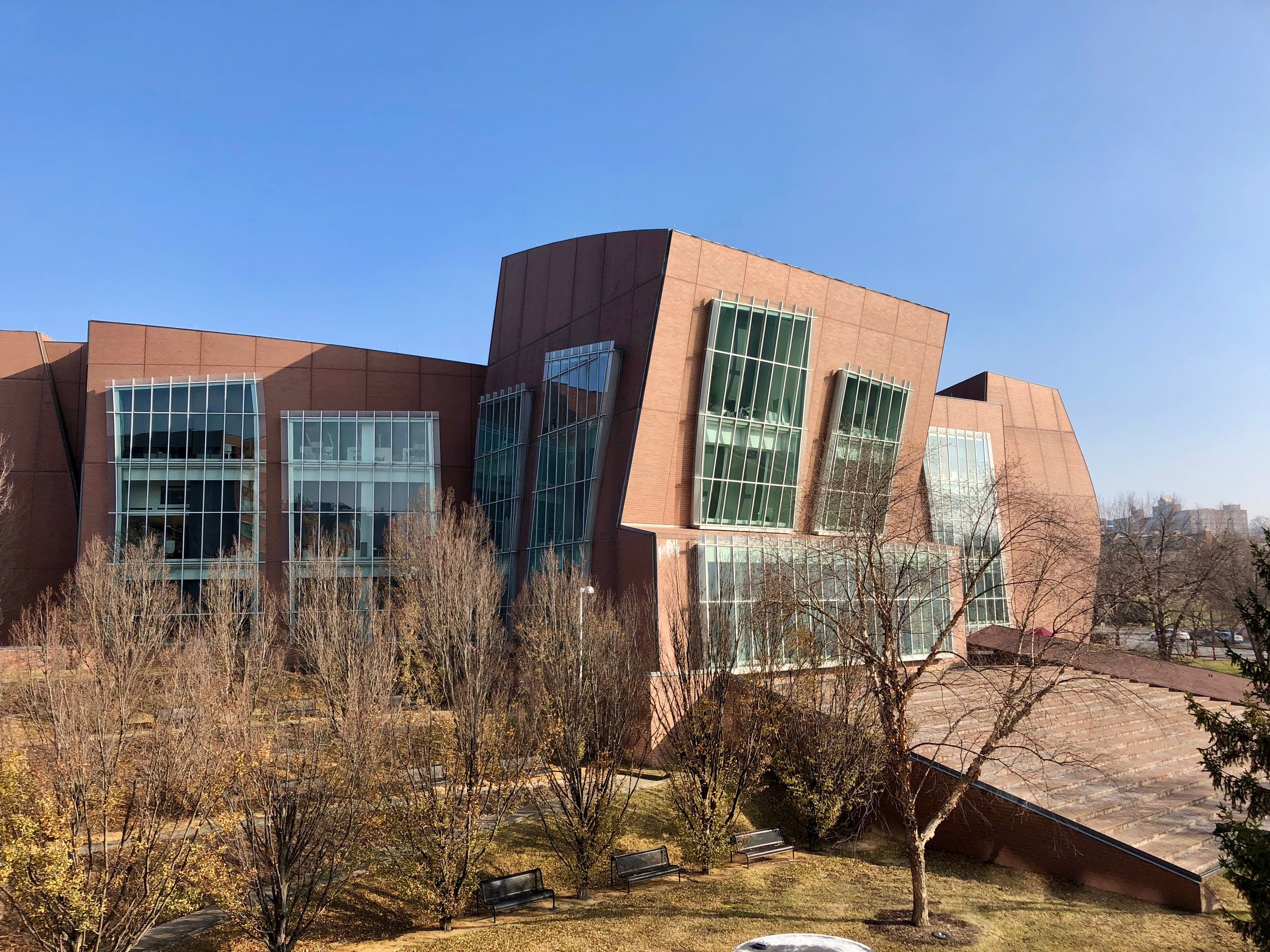 Großes Gebäude mit vielen Fenstern, umgeben von Bäumen, Bänken, Gras und einem Laternenmast, mit anderen Gebäuden und einem klaren blauen Himmel im Hintergrund; als der neue Campus der University of Illinois at Chicago bezeichnet.