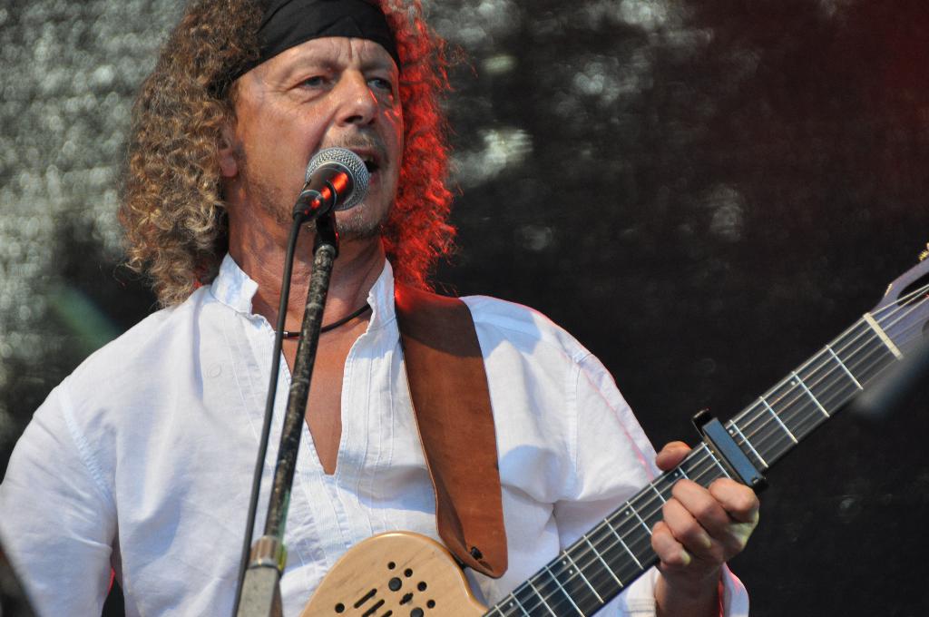 Mann in weißem Hemd und schwarzem Stirnband singt in ein Mikrofon und hält eine Gitarre