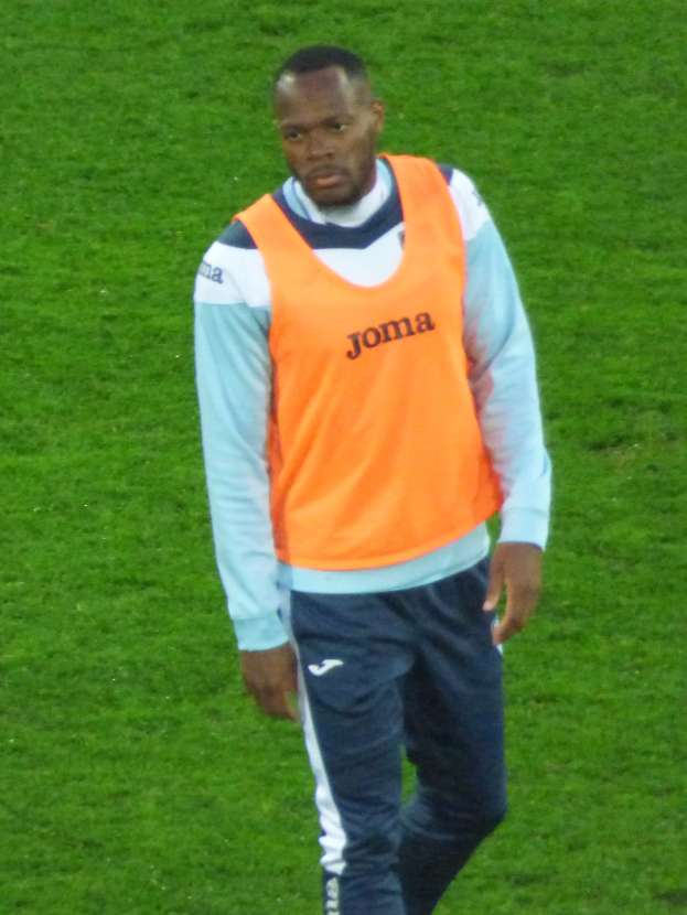 Ein Fußballspieler in einem orangen T-Shirt und blauen Hosen läuft entschlossen über ein grünes Feld.