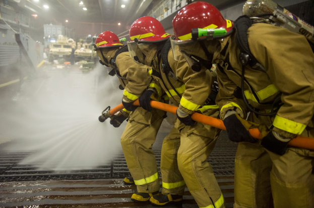 Feuerwehrleute in Helmen und Handschuhen halten Schläuche und spritzen Wasser auf ein Feuerwehrauto, mit verschiedenen Gegenständen und einem Boden im Hintergrund.