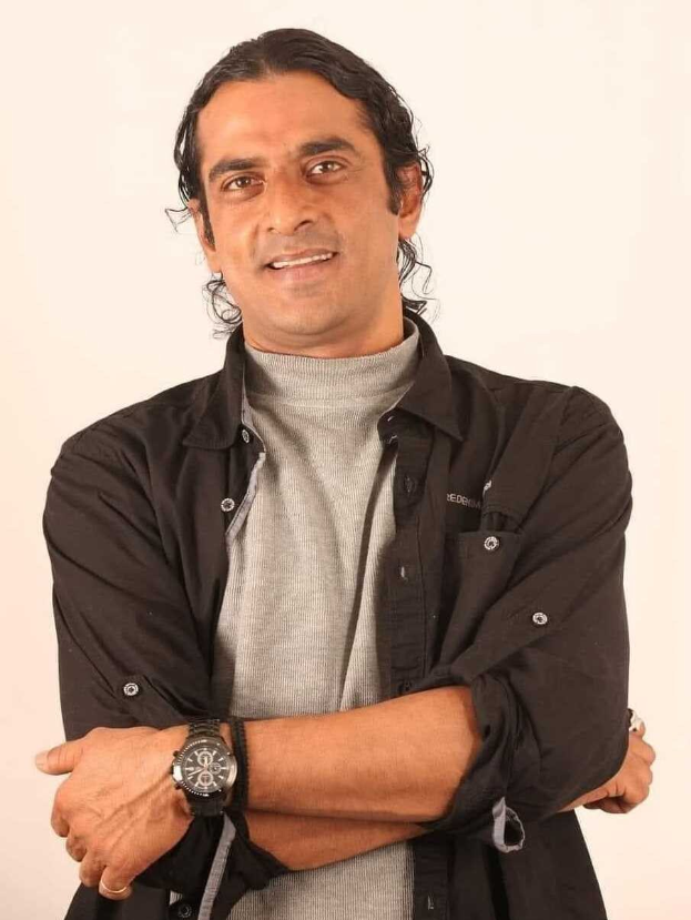 Ein Mann in einem schwarzen und aschfarbenen Kleid mit überkreuzten Armen, der eine Uhr trägt, vor einem weißen Hintergrund steht und nachdenklich scheint.