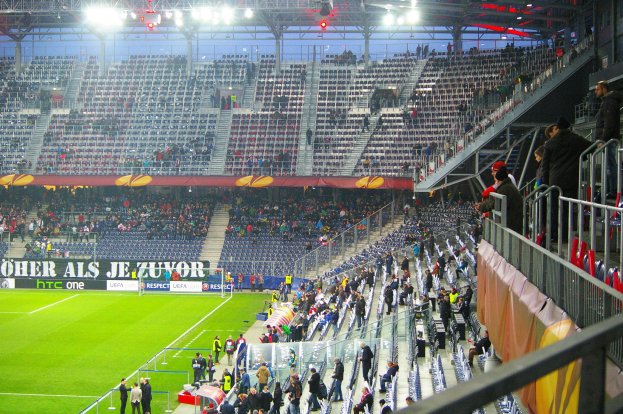 Ein großes Stadion mit Zuschauern, die ein Fußballspiel anschauen, mit einigen Menschen, die auf Stühlen stehen oder sitzen, einem Geländer und einer Fahne auf der rechten Seite und einem leeren Stadion mit Deckenbeleuchtung im Hintergrund.