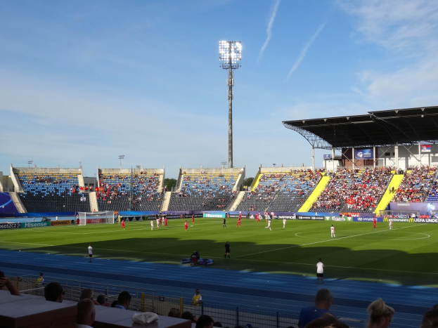 Ein Fuballspiel in einem gro├čen Stadion mit Spielern auf dem Feld und Zuschauern in den R├Ąngen unter Flutlicht.