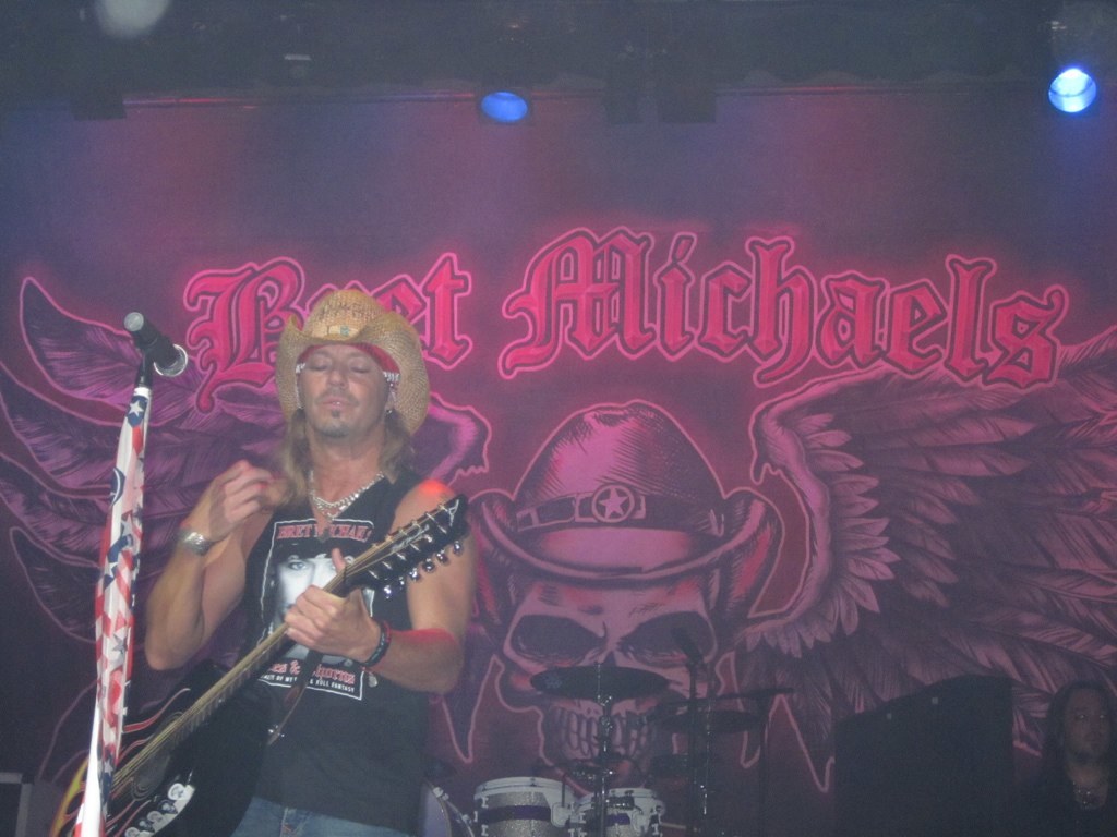 Eine Musikkonzertszene mit einem Gitarristen links, einer Person in der rechten unteren Ecke, einem Banner im Hintergrund und Drogen in der Mitte unten.