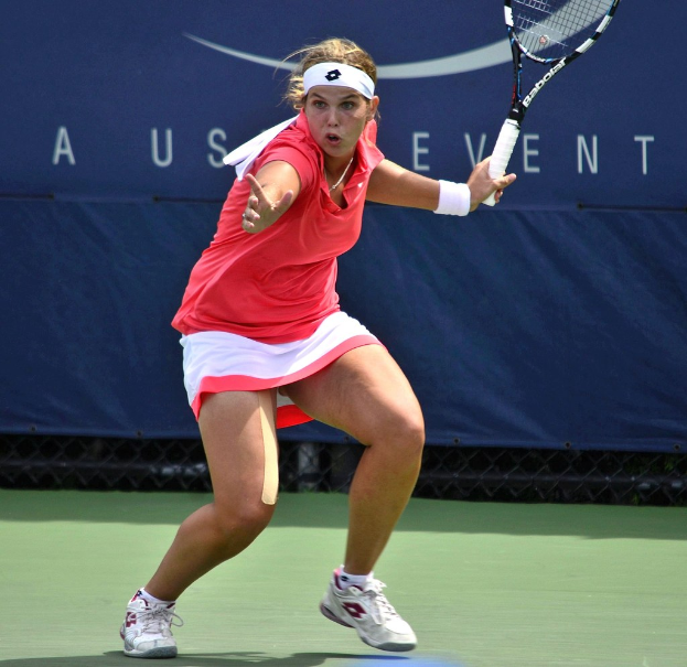 Eine Frau in einem pinken Shirt und weißen Schuhen, mit einem Headband, spielt Badminton auf einem Court, hält einen Schläger und hat ein Banner im Hintergrund.