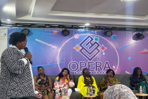 Eine Frau, die Gründerin des Opera Event Space, steht mit einem Mikrofon vor einer Gruppe sitzender Frauen, mit Flaschen und anderen Gegenständen auf den Tischen im Vordergrund und einem Banner mit Text und Deckenbeleuchtung im Hintergrund.