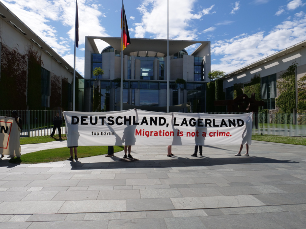 Eine Gruppe von Menschen, die ein Transparent mit der Aufschrift "Deutschland, Lagerland Migration ist kein Verbrechen" vor einem eingezäunten Gebäude mit Bäumen und Flaggen hält.
