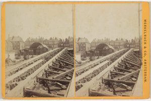 Ein altes Schwarz-Weiß-Foto einer Bahnschiene mit Gebäuden im Hintergrund, Text auf der rechten Seite lesbar "Merkelbach & Co. Amsterdam" und verstreute hölzerne Gegenstände im Vordergrund.