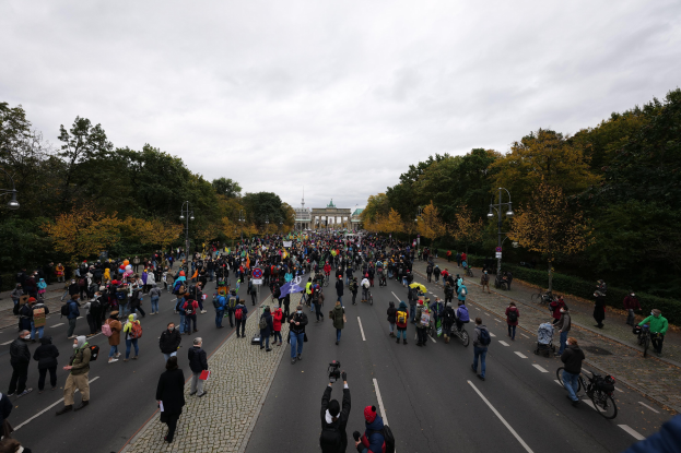 Eine große Gruppe von Menschen, die eine von Bäumen gesäumte Straße in Berlin entlanggehen und Kameras halten, mit einem Gebäude und einem klaren Himmel im Hintergrund.