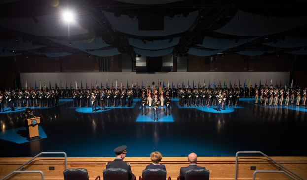 Große Gruppe uniformierter Soldaten auf einer Bühne mit einem Podium und Mikrofon, während sitzende Personen unten beobachten und eine beleuchtete Wand den Hintergrund bildet während einer Militärzeremonie.