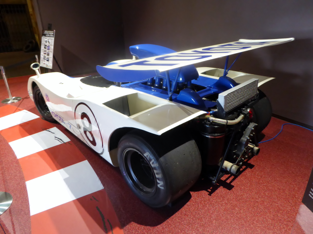 Ein weißer und blauer Rennwagen in einem Museum ausgestellt, umgeben von Absperrbögen und Schautafeln, mit einer Wand im Hintergrund, die auf das Goodwood Festival of Speed hinweist.