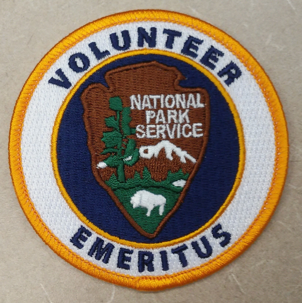 Runder Aufnäher mit "Volunteer National Park Service Emeritus" in fetter schwarzer Schrift auf weißem Hintergrund, umgeben von einem gelben Kreis und einem blauen Rand.