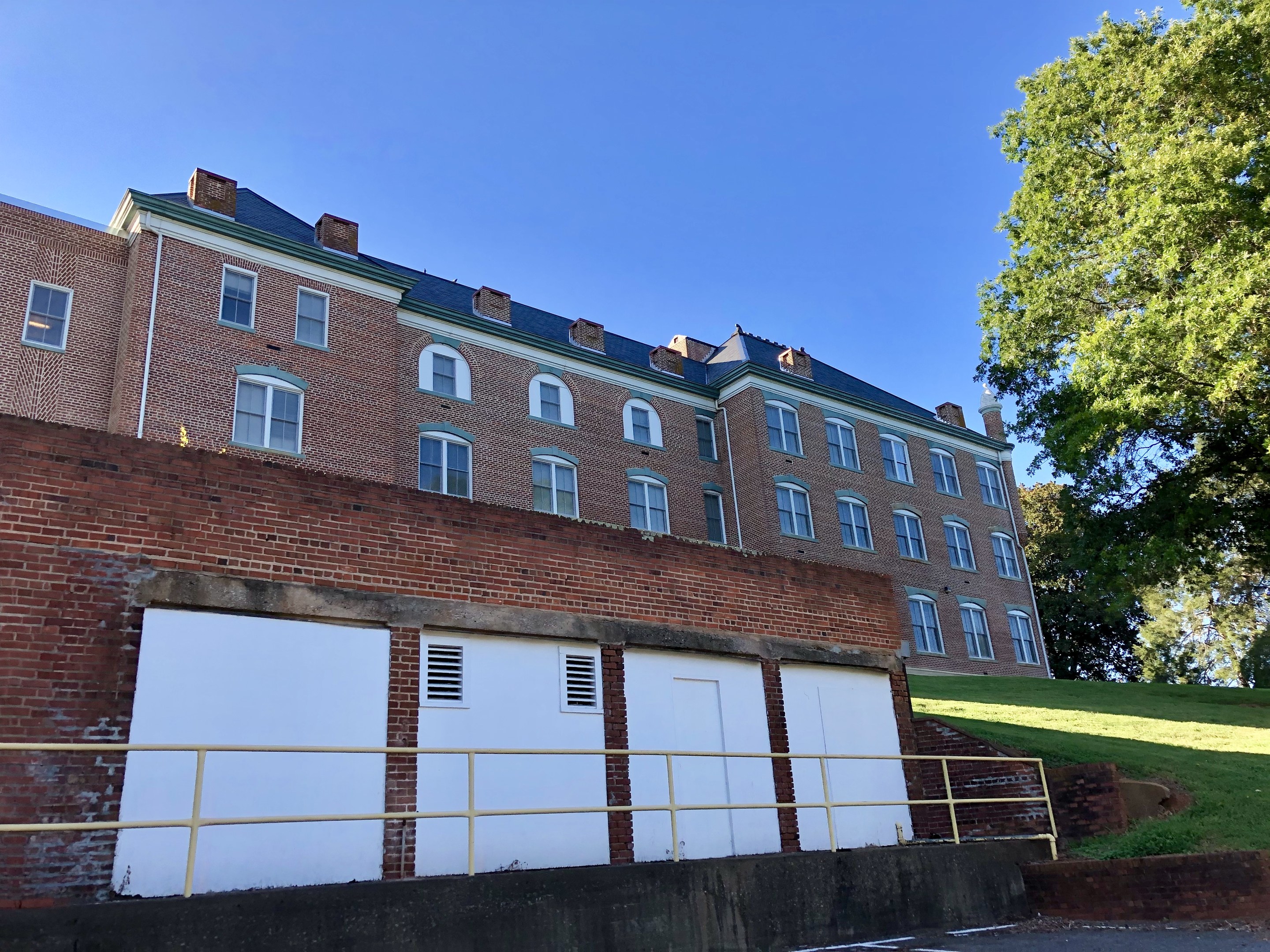 Großes Backsteingebäude mit zahlreichen Fenstern, umgeben von einem Metallzaun, Weg, Gras, Bäumen und einem bewölkten Himmel, auf dem Campus der University of Virginia gelegen.