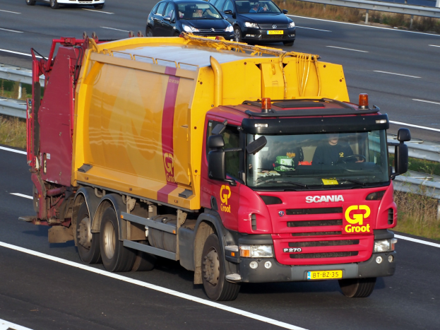 Ein gelber und roter Müllwagen mit einer Begrenzung auf einer Autobahn, umgeben von anderen Autos und Grünflächen.