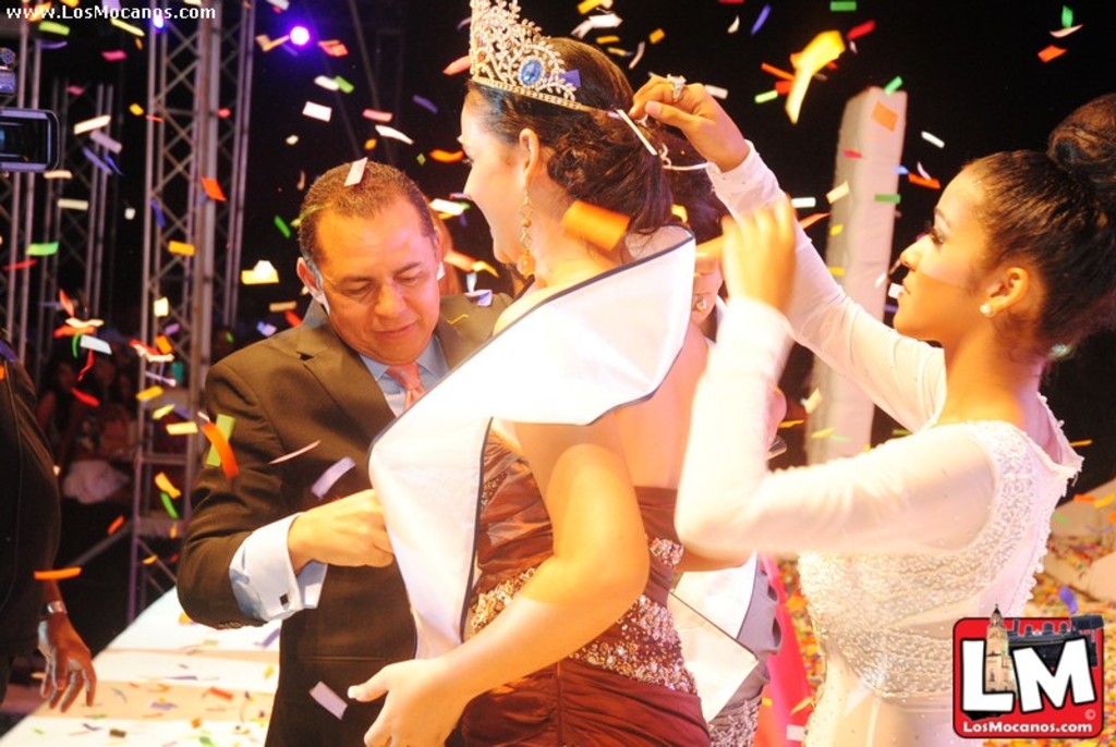 Ein Mädchen mit einer Krone steht auf einer Bühne bei einer Miss World Show, während eine andere Frau die Krone hinter ihr anpasst und ein Mann in einem schwarzen Anzug vor ihr die Hand schüttelt, vor einem Hintergrund aus Metallrahmen und einer dunklen Kulisse.