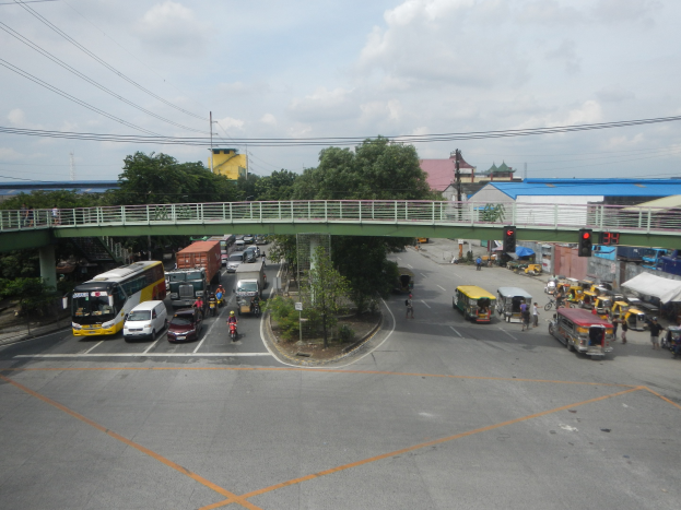 Vollgestaute Stadtstraße mit Autos, Bussen, Fußgängern, Brücke, Ampeln, Gebäuden, Bäumen, Strommasten und bewölktem Himmel.