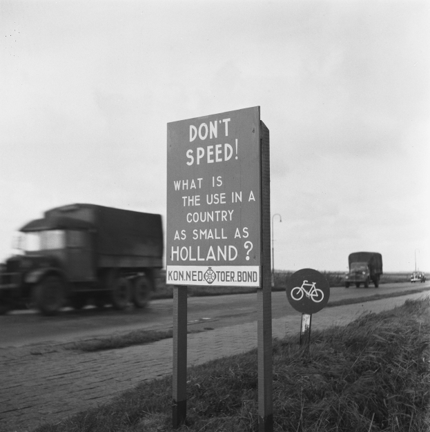 Schwarz-weißes Foto eines Lkw auf einer Straße mit einem "Fahren Sie nicht zu schnell"-Schild im Vordergrund und anderen Fahrzeugen, Gras, Pfosten und einem klaren Himmel im Hintergrund.