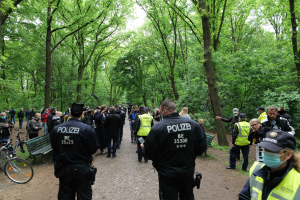 Polizeibeamte vor einer Menschenmenge bei einem Outdoor-Event mit Fahrrädern und einer Bank im Vordergrund und Bäumen im Hintergrund.