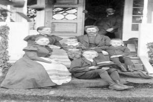 Ein Schwarz-Weiß-Foto einer Familie von fünf Personen, die auf der Treppe eines Hauses sitzen, mit einem Mann im Hintergrund, das Haus hat eine Tür und Fenster.