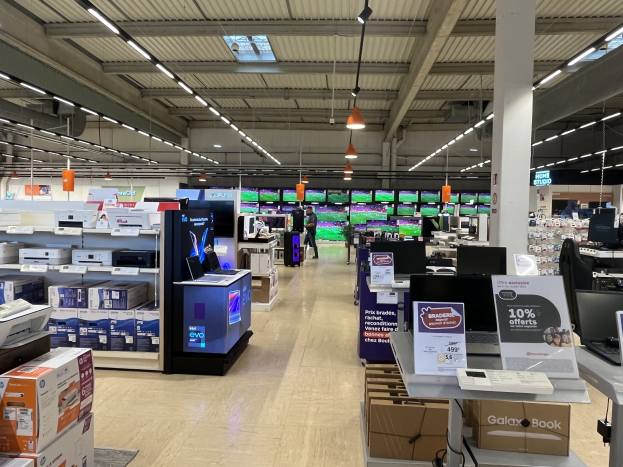 Großer Raum mit Elektronikgeräten einschließlich Computern, Druckern und Kisten, die in Regalen angeordnet sind, Menschen auf dem Boden, eine Säule, Schilder, Deckenleuchten und Deckenventilatoren in einem kürzlich eröffneten Walmart-Laden.