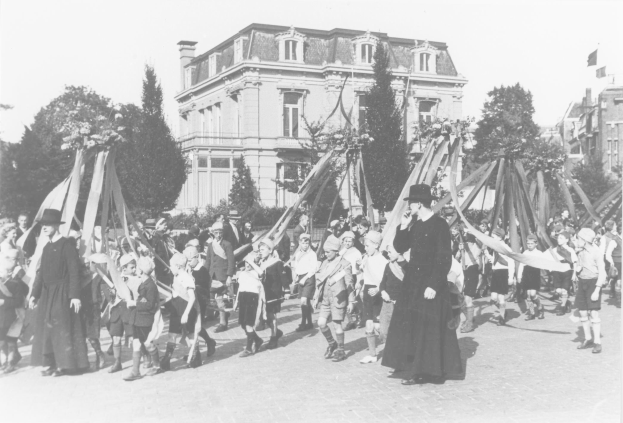 Schwarz-weißes Bild einer Gruppe von Menschen, die die Straße entlanggehen und Fahnen schwenken, mit Hüten, Bäumen und Gebäuden im Hintergrund, mit einem Maibaum als Symbol für den Maifeiertag.