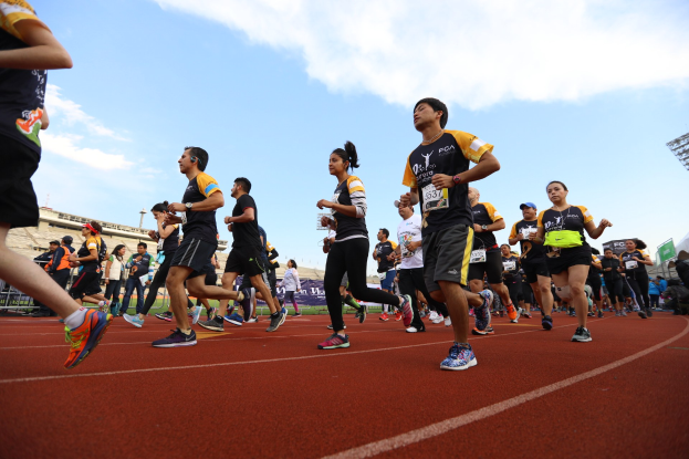 Gruppe von Menschen, die auf einer Laufbahn laufen, mit Zuschauern auf der rechten Seite, Gebäuden und Bannern im Hintergrund unter einem klaren blauen Himmel.