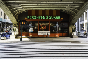 Eine belebte Stadtkulisse mit einem zentralen Gebäude, einem Schild mit der Aufschrift "Pershing Square Cafe in New York City", abgestellten Fahrrädern, Topfpflanzen, einer Straßenlaterne, einem Metallzaun, Menschen auf Bänken sitzend, Bäumen und einem bewölkten Himmel.
