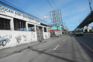 Eine Straße in einer Stadt mit Fahrzeugen, eine Brücke auf der rechten Seite, Gebäude, Strommästele mit Kabeln und einen bewölkten Himmel; ein mit Graffiti beschmiertes Gebäude ist an der Straßenseite zu sehen.