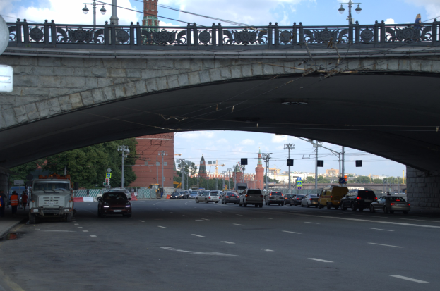 Eine Brücke überspannt eine vielbefahrene Straße mit darunterfahrenden Fahrzeugen, während Fußgänger auf dem Gehweg stehen; Gebäude, Bäume, Verkehrsampeln und Laternen sind im Hintergrund zu sehen.