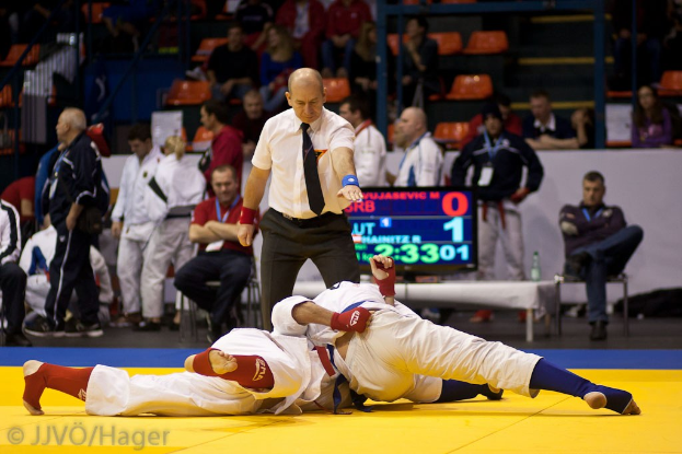 Zwei Judoka im Kampf mit einem Schiedsrichter in der Nähe, während Zuschauer im Hintergrund zusehen.
