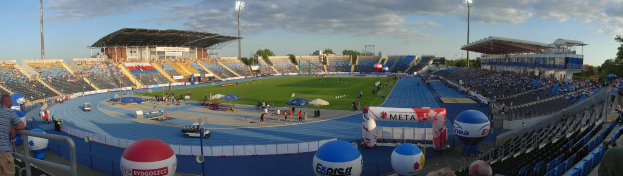Großes Stadion mit Zuschauern bei einer Leichtathletikveranstaltung, umgeben von Bäumen, Laternenmasten und Gebäuden unter einem bewölkten Himmel.