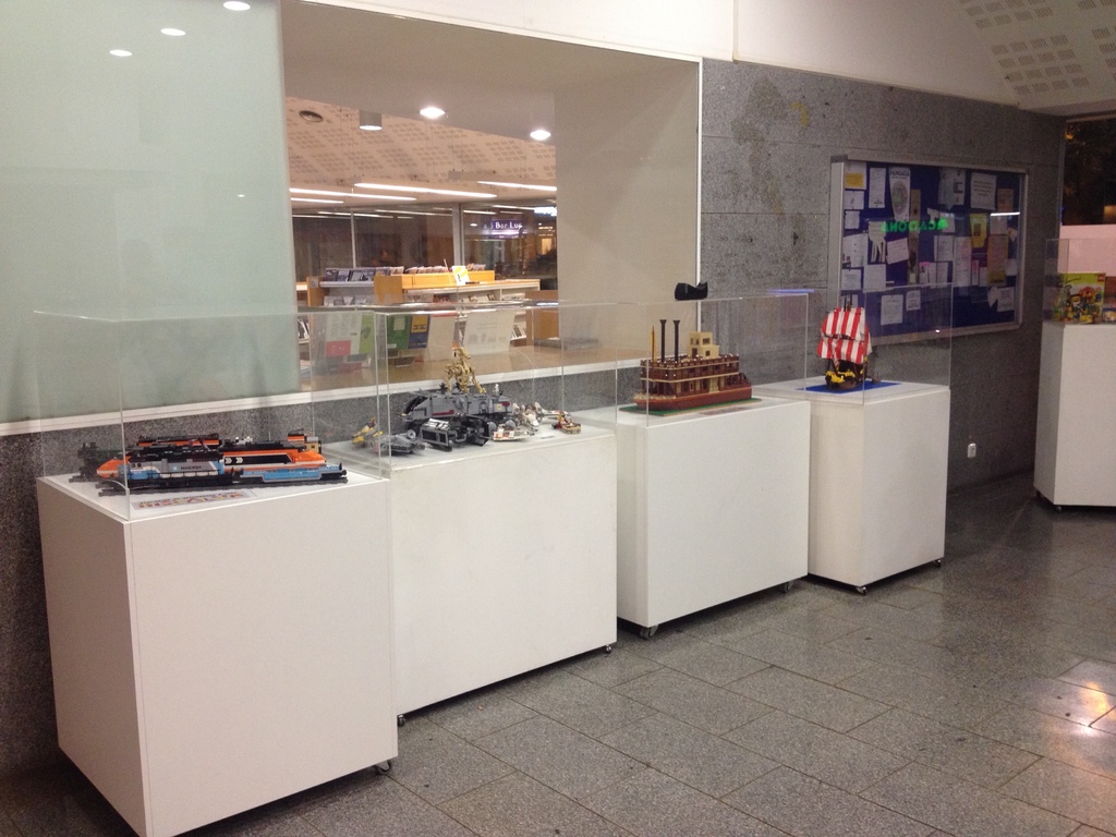 Artikel, Die Auf Weißen Tischen In Einer Ausstellungshalle Ausgestellt Sind, Mit Einer Ankündigungstafel, Die Mit Plakaten Belegt Ist, An Der Wand Dahinter Und Einer Decke, Die Oben Im Bild Sichtbar Ist.